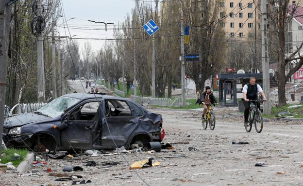 Two cyclists pedal down a street in Mariupol where traces of war are visible.